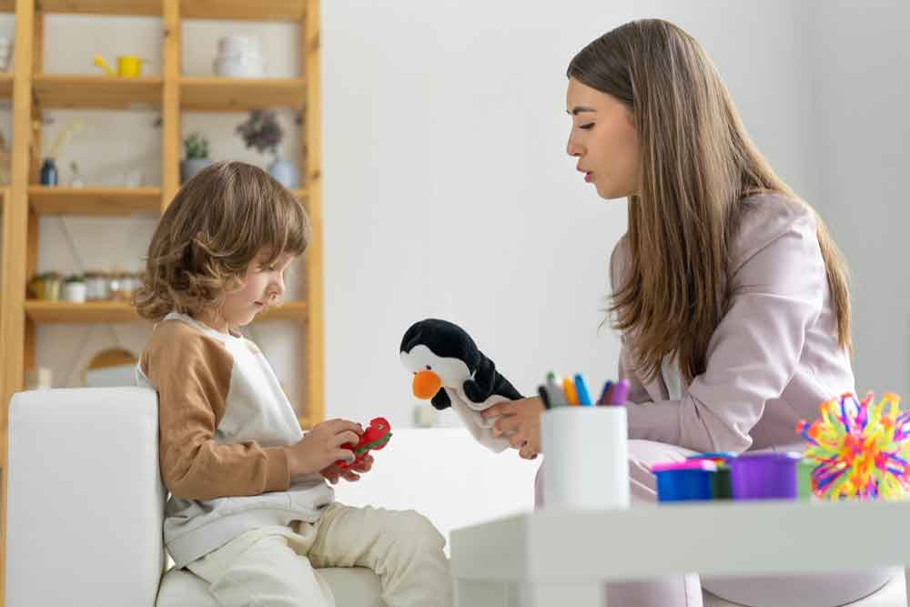 Young woman psychologist plays with her little client. Holds penguin doll in her hand. The boy is playing with dragon toy.